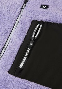 Giacca in pile viola con tasca zip nera, cerniere robuste e superficie morbida e strutturata. Etichetta del brand visibile sul tessuto.