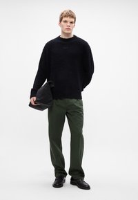 Pulover din tricot negru texturat cu guler rotund, asociat cu pantaloni verzi închis. Modelul ține o geantă neagră, purtând pantofi din piele negri.