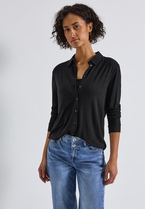 Camicetta nera con colletto, texture morbida e maniche lunghe, abbinata a jeans di denim azzurro chiaro con dettagli dei bottoni in vista.