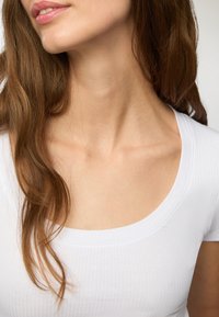 T-shirt bianca a costine con scollatura a girocollo; tessuto morbido e maniche corte; capelli castani leggermente ondulati che ricadono di lato.