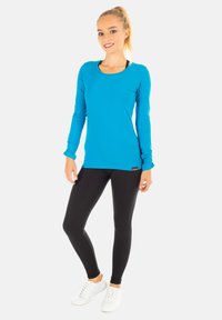 Langärmliges türkisfarbenes Sportoberteil mit runder Ausschnitt, kombiniert mit schwarzen Leggings. Glattes Material und taillierte Passform, geeignet für aktiven Lebensstil.