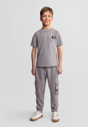 Chłopiec stojący, ubrany w szarą koszulkę z krótkim rękawem i pasujące spodnie bojówki z kieszeniami oraz białe trampki na jednolitym tle.