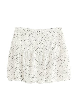 Smilende pige står iført en sort tanktop, hvid fluffy cardigan, hvid nederdel med polkaprikker og beige sneakers på en hvid baggrund.