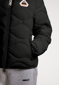Giacca nera imbottita con design trapuntato, dotata di cerniera frontale e tasche laterali. Abbinata a pantaloni da tuta grigi con la scritta "JACKIT" in nero.