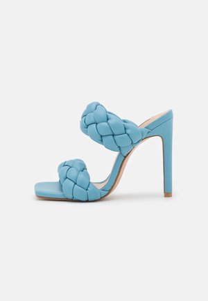 Blå højhælet sandal med firkantet tå og to brede flettede remme hen over foden og tæerne, vist mod en ensfarvet hvid baggrund.