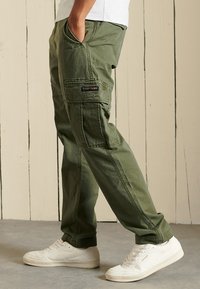 Olivengrøn cargo-bukser med flere lommer, en afslappet pasform og forstærkede sømme, parret med hvide sneakers mod en neutral baggrund.