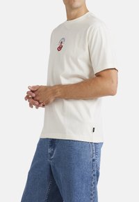 Camiseta de algodón de manga corta en color crema, con un diseño gráfico rojo y acentos azules. Combinada con jeans de mezclilla azules.