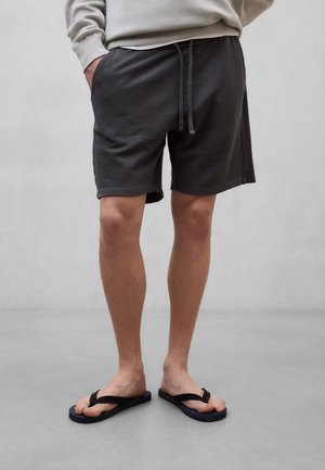 Mand iført sorte afslappede shorts med snor og sorte klipklappere, stående mod en ensartet lys grå baggrund.