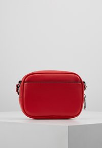 Malá obdélníková červená kožená crossbody taška se zipovým uzávěrem a nastavitelnými bočními popruhy, umístěná na bílé podešvi.