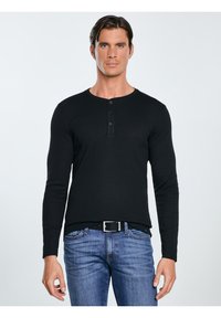 Mężczyzna ubrany w czarną koszulę henley z długim rękawem i niebieskie dżinsy z czarnym paskiem, stojący na tle jednolitej białej ściany.