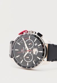 Μαύρο ρολόι χειρός χρονογράφου με στεφάνη ταχύμετρου, τρεις υποκαντράν, παράθυρο ημερομηνίας στη θέση 3 και μαύρο δερμάτινο λουράκι.