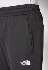 Černé sportovní kalhoty se strukturovaným povrchem, vybavené elastickým pasem a boční kapsou. Na nich je bíle vyšitá značka The North Face.