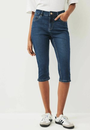 Pantalons capri en denim bleu foncé, à coupe ajustée, avec cinq poches, ourlet retroussé et détails de couture contrastants. Portés avec des baskets blanches.