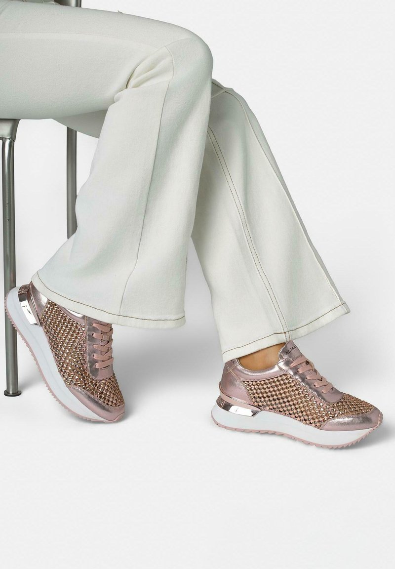 Persona che indossa pantaloni svasati di colore chiaro e scarpe da ginnastica metallizzate rosa oro con dettagli in strass, seduta su uno sgabello contro uno sfondo bianco.