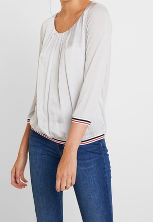 Woman wearing white pleated blouse with striped cuffs and hem, paired with blue denim jeans, standing against a plain white background.