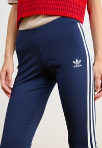 Sötétkék leggings fehér oldalsávokkal. Simább, rugalmas anyagból készült. Az Adidas logója a bal comb felső részén található.