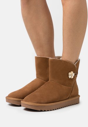 Brown suede ankle boots with a rounded toe, shearling lining, and decorative floral hardware detail on the side. Ribbed rubber soles.