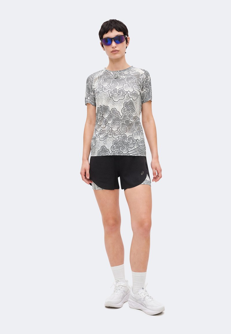Person iført sort og hvid mønstret T-shirt, sorte sportsshorts, hvide sokker, hvide sneakers og blå sports solbriller, står foran en ensfarvet baggrund.