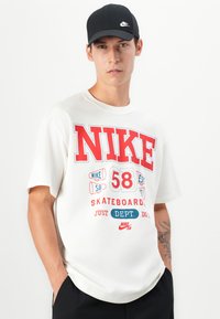 White cotton T-shirt with large red "NIKE" branding and playful graphics including "58," "SKATEBOARD," and "JUST DO IT." Short sleeves, relaxed fit.