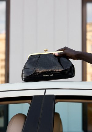 Main à la peau foncée tenant une pochette Valentino noire à texture crocodile au-dessus d’un toit de voiture blanc.