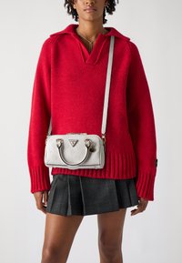Maglione rosso lavorato a maglia con scollatura a V e polsini a coste, abbinato a una borsa bianca con texture dotata di manico superiore e lunga tracolla.
