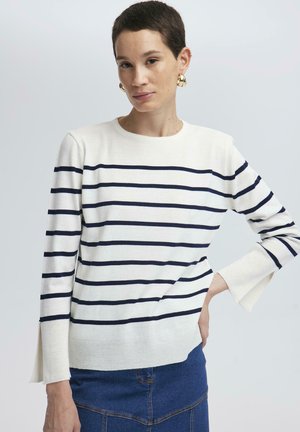 Femeie cu păr scurt, purtând un pulover alb cu dungi orizontale bleumarin și cercei aurii tip cerc, asortat cu o fustă de denim.