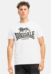 Balta medvilninė T-shirt maikė su dideliu juodu liūto logotipu ir „LONSDALE LONDON“ tekstu sodria juoda šriftu per krūtinę. Trumpomis rankovėmis.