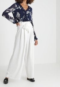 Blusa floreale blu marina in tessuto trasparente con scollatura arricciata, abbinata a pantaloni bianchi a gamba larga e scarpe flat nere.
