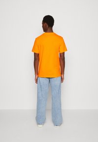 Hombre de espaldas vistiendo una camiseta de manga corta de color naranja brillante, jeans holgados de color azul claro y zapatillas blancas frente a una pared gris lisa.