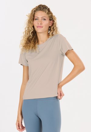 Vrouw met krullend blond haar die een beige shirt met korte mouwen en blauwe leggings draagt, staande met één hand op haar heup tegen een witte achtergrond.