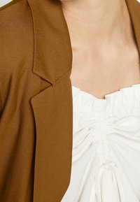 Brun kavaj med strukturerade revers, matchad med en vit blus med rynkdetalj och dragsko. Slät tygtextur, elegant design.