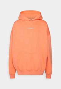 Överdimensionerad hoodie i stark orange tyg, med en framficka och vit grafisk text "© WORST BEHAVIOR" på bröstet.