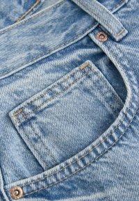 Tissu en denim bleu clair avec une poche avant et des coutures visibles. La texture est légèrement délavée avec une finition lisse et du matériel métallique.