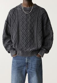 Pull en maille gris foncé avec motifs torsadés texturés, manches larges et poignets et ourlet côtelés. Associé à un jean en denim bleu clair.