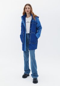 Giacca impermeabile blu con cappuccio, tasche frontali con piping bianco a contrasto, abbinata a jeans svasati azzurri chiari e scarpe nere.