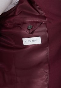 Interior de chaqueta burdeos con un forro suave y brillante, cierre de un solo botón y una etiqueta blanca que dice "PIER ONE."