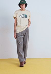 Beige t-shirt med grøn logo, grå kordfløjspants og brune sneakers. Modellen står mod en lyseblå væg på et gult gulv.