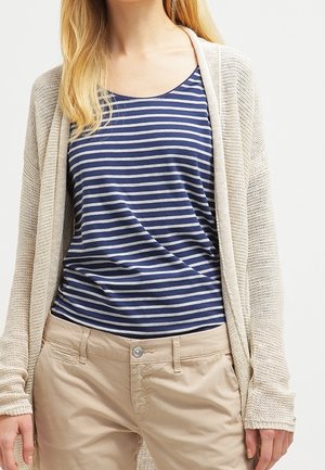 Frau trägt ein blau-grau gestreiftes ärmelloses Oberteil, eine beige Strickjacke mit offenem Muster und hellbraune Hose, steht vor einem weißen Hintergrund.