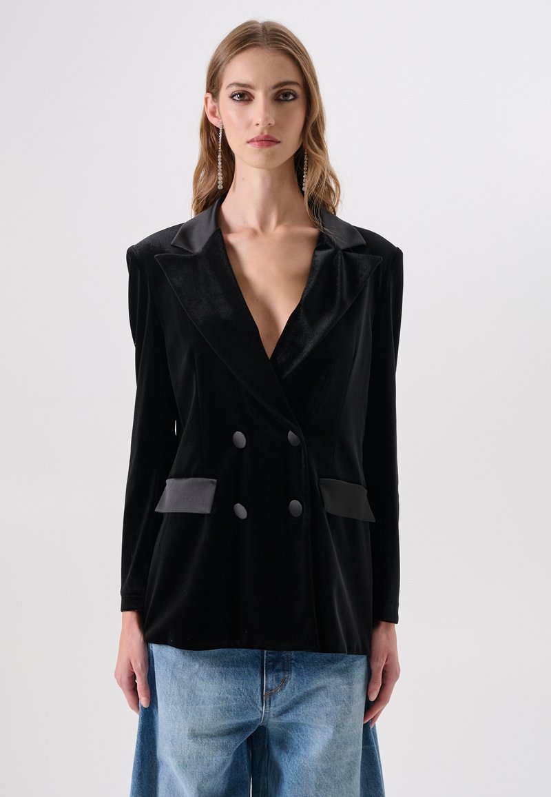 Zwarte fluwelen dubbelgeknoopt colbert met satijnen revers, voorzien van twee voorzakken en grote knopen, gedragen over losse blauwe denim.