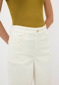 Vita denimbyxor med hög midja och en slät yta, femfickorsdesign och guldknappsstängning. Ribbade olivgröna toppen kompletterar outfiten.