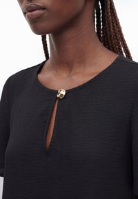 Femme aux cheveux tressés portant un haut noir texturé fermé par un seul bouton rond doré au col.