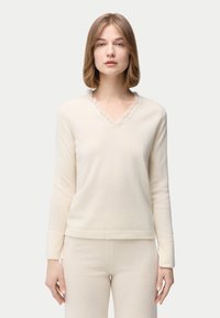 Cremefarbener Pullover mit V-Ausschnitt und Rüschenbesatz, aus weichem Stoff gefertigt. Lange Ärmel und gerader Saum sorgen für eine entspannte Passform.