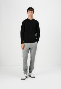 Suéter de punto negro, pantalones de rayas gris claro, zapatillas blancas con rayas negras. Gafas de sol en la cabeza. Pose de pie contra un fondo blanco.