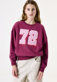 Sudadera burdeos con un gran número "72" texturizado en rosa y contorno rojo. Cuello y puños de canalé, corte relajado, combinada con pantalones claros.