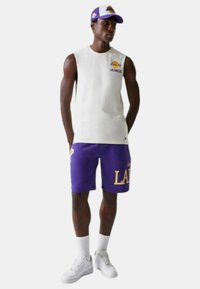 Maglietta bianca senza maniche con il logo "Lakers Los Angeles", abbinata a pantaloni corti viola con "LAK" in giallo. Sneakers bianche e cappellino viola e bianco.