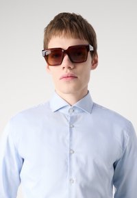 Brown oversized sunglasses with a geometric design and glossy finish. Worn with a light blue, long-sleeved, button-up shirt.