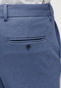 Μπλε υφασμάτινο παντελόνι με μεγάλη πίσω τσέπη και μαύρη λεπτομέρεια κουμπιού. Το ύφασμα παρουσιάζει υφαντό μοτίβο με διακριτική λάμψη.