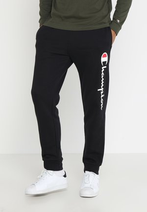 Mężczyzna w czarnych spodniach dresowych z białym i czerwonym logo Champion na lewej nodze oraz białych tenisówkach, stoi z rękami w kieszeniach.