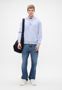 Svetlomodrá mikina s kapucňou a prednými vreckami, zdobená centrálne vyšitým logom. Modré zvonové džínsy a biele tenisky.