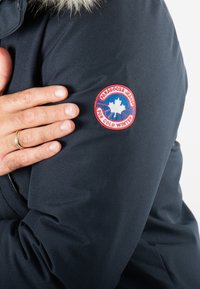 Parka d'hiver marine avec un col en laine, affichant un patch rond proéminent avec une bordure rouge, des lettres bleues et un motif de feuille d'érable.